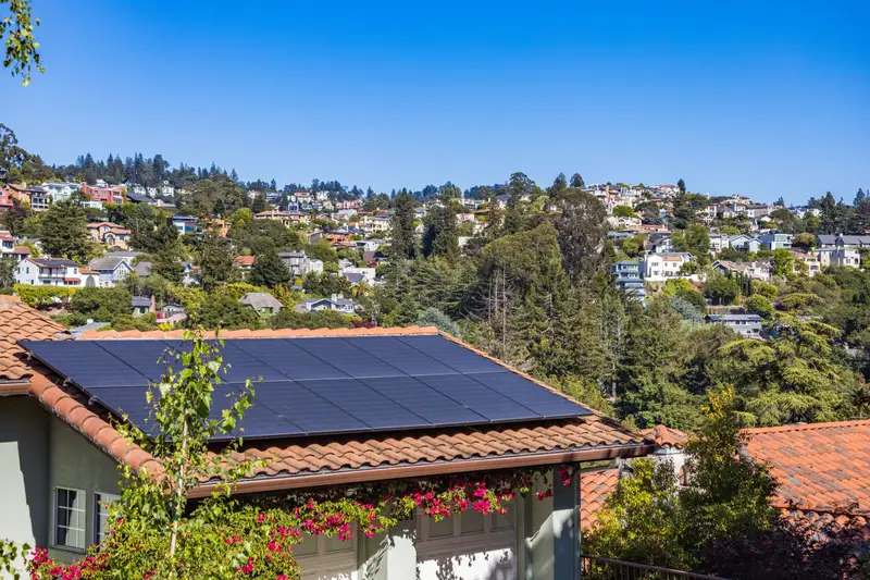 Maison au toit de tuiles couvert de panneaux solaires noirs, en plein soleil sous un ciel bleu, entourée de verdure
